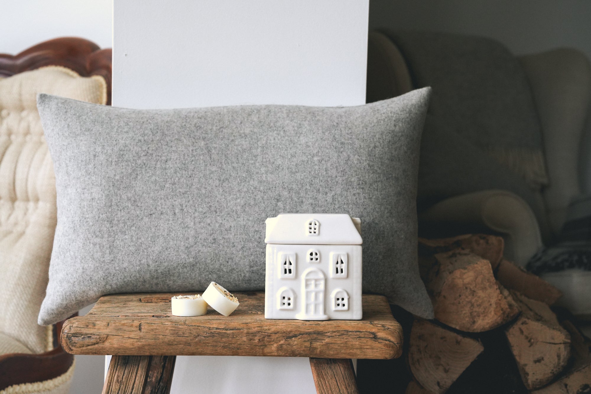 A rectangular grey throw pillow displayed on a rustic wooden stool in a cosy living room setting, with a small white cottage wax burner -with two cashmere botanical soy wax melts next to it.