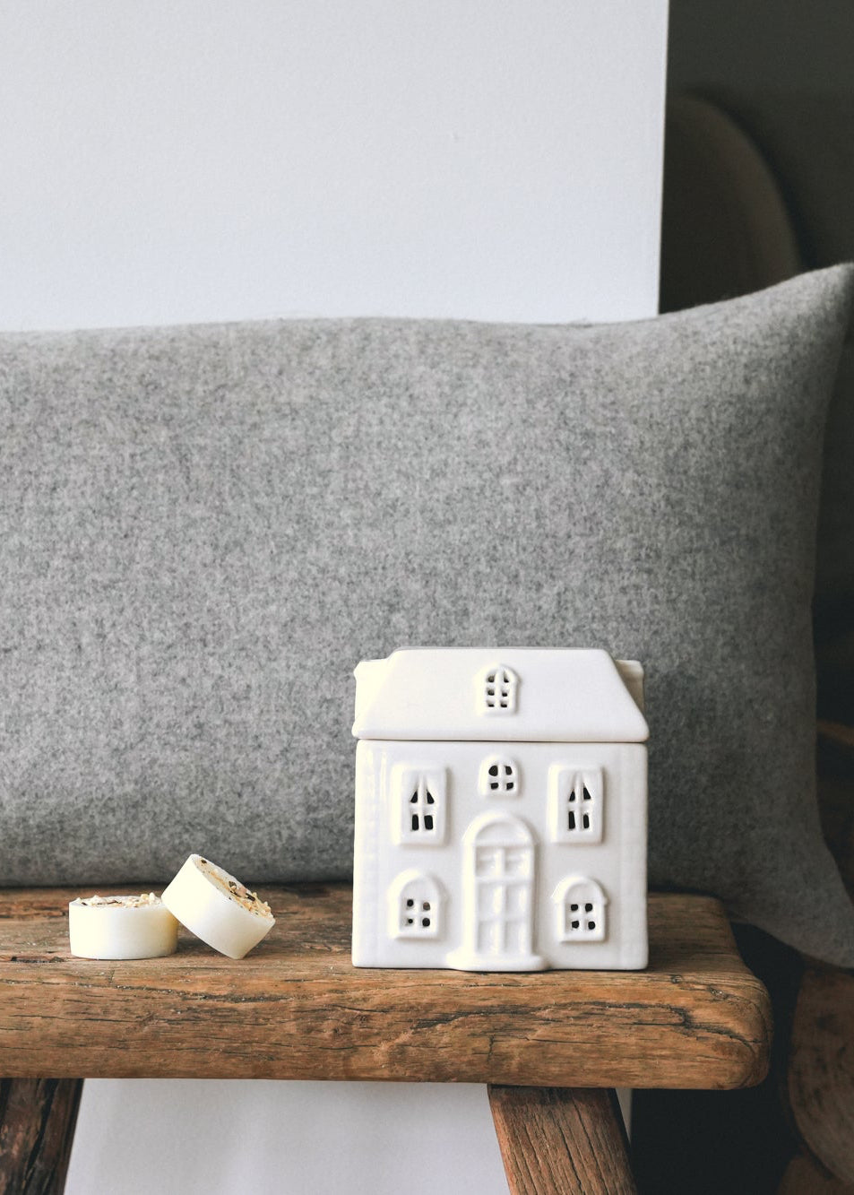 A rectangular grey throw pillow displayed on a rustic wooden stool in a cosy living room setting, with a small white cottage wax burner -with two cashmere botanical soy wax melts next to it.