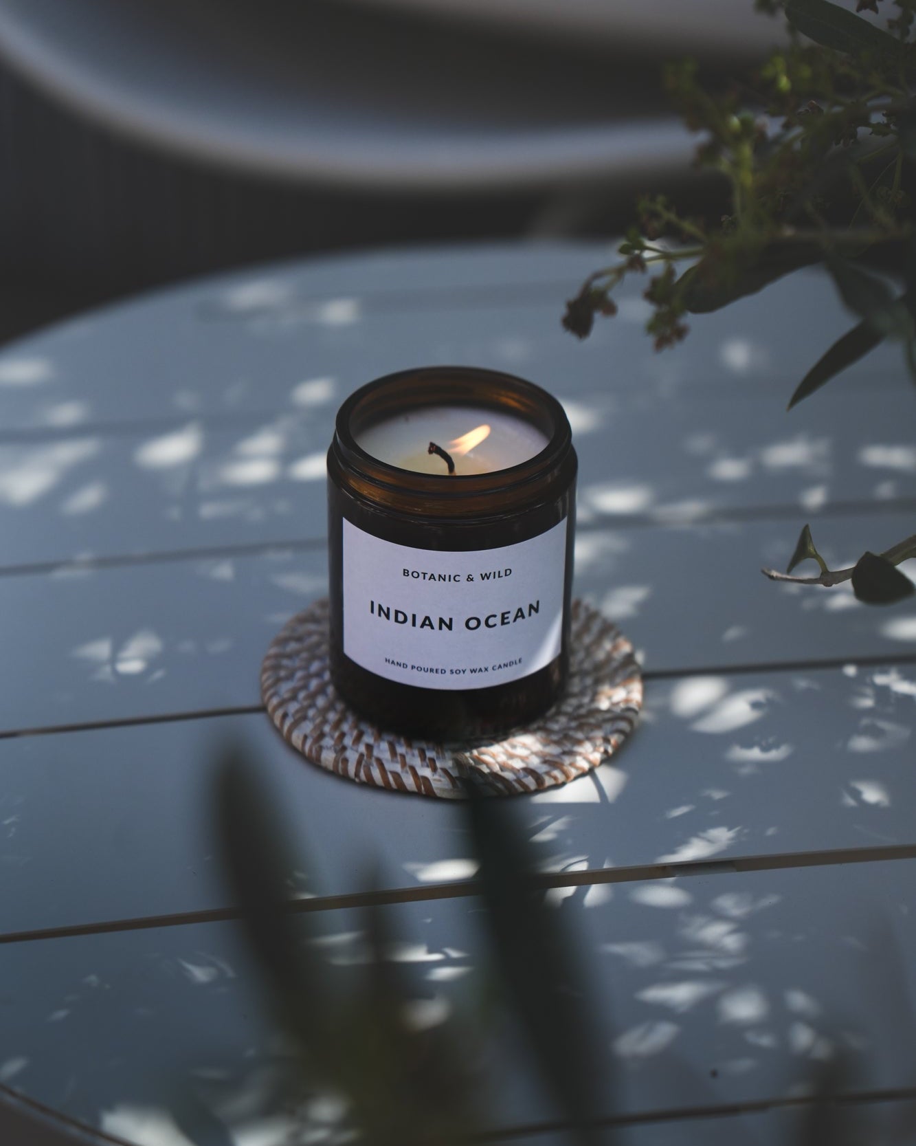 A lit amber glass candle jar labeled 'INDIAN OCEAN' sits on a woven coaster on a light blue wooden table, with soft dappled sunlight and blurred greenery in the foreground and background.