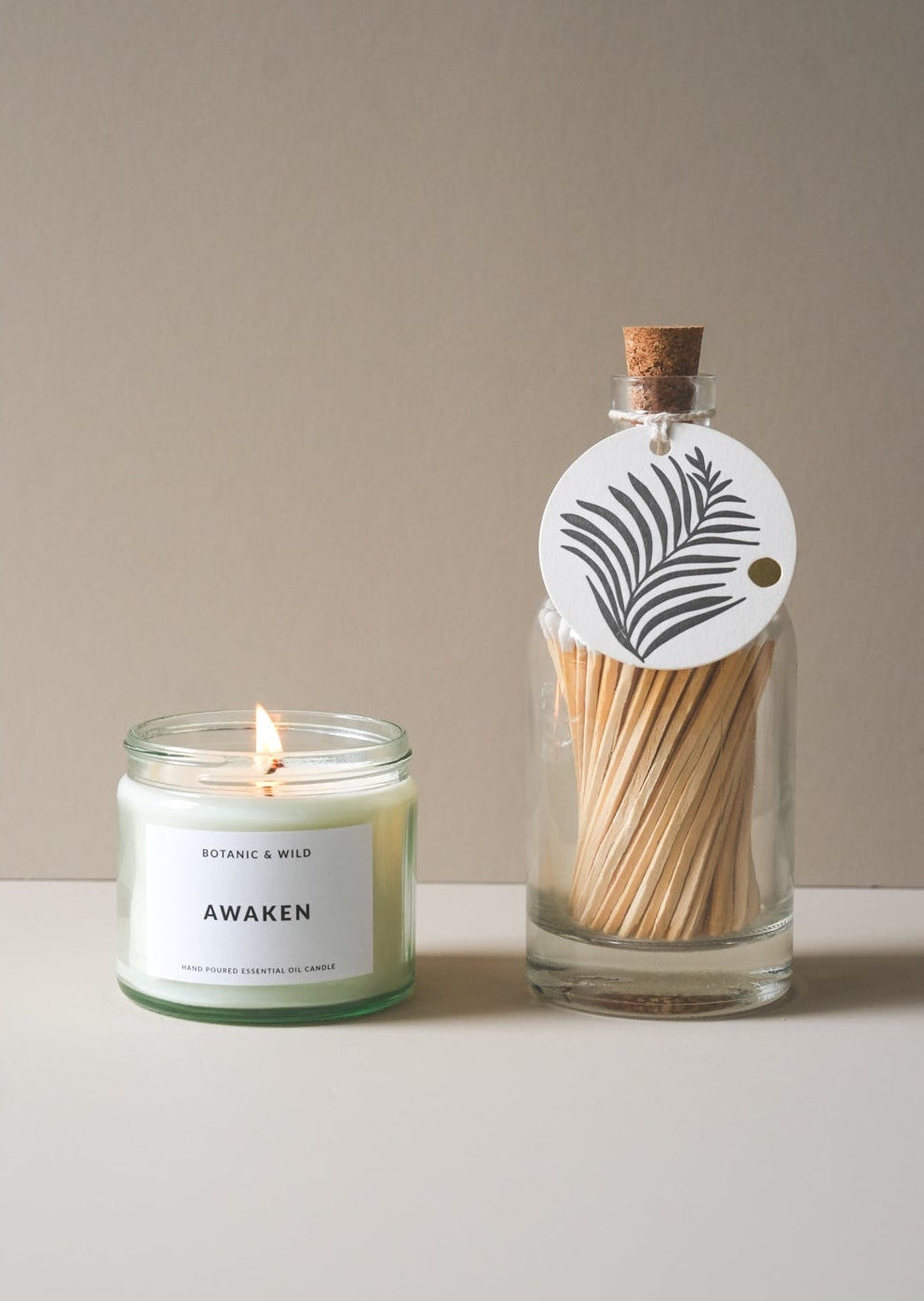 A lit candle in a clear glass jar with a white label reading 'AWAKEN' sits beside a clear glass bottle filled with wooden matches and a hanging white tag with a palm leaf illustration, set against a warm beige background.