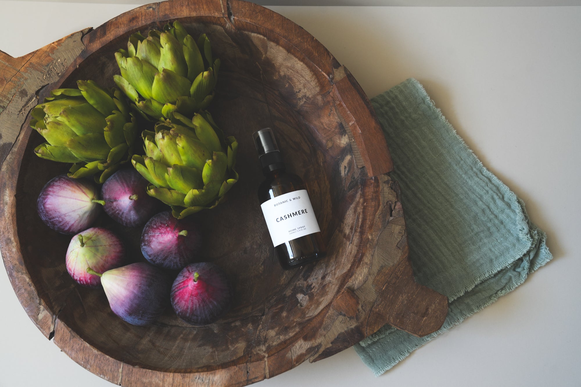A small dark glass bottle home spray with a white label reading CASHMERE, styled in a rustic wooden bowl with green artichokes and purple figs, next to a folded sage green cloth on a light neutral surface.